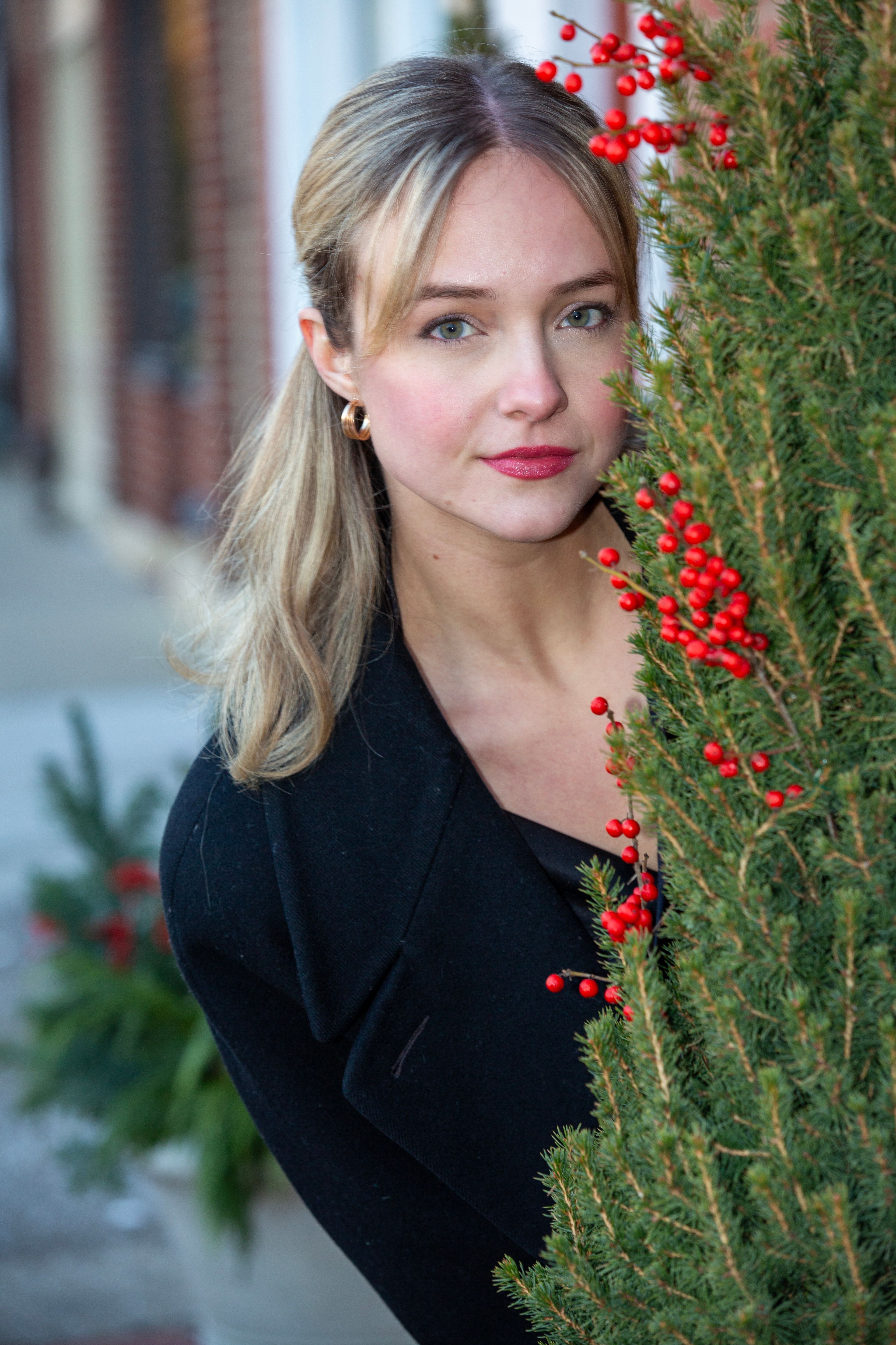 Young woman playfully peeking behind a festive evergreen with red berries, outdoors.