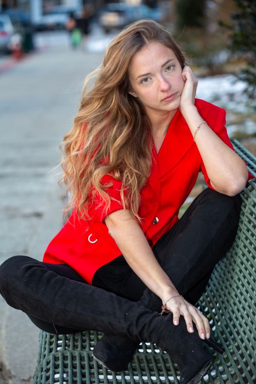 Free Portrait of a thoughtful young woman in a vibrant red jacket sitting on a bench outdoors. Stock Photo