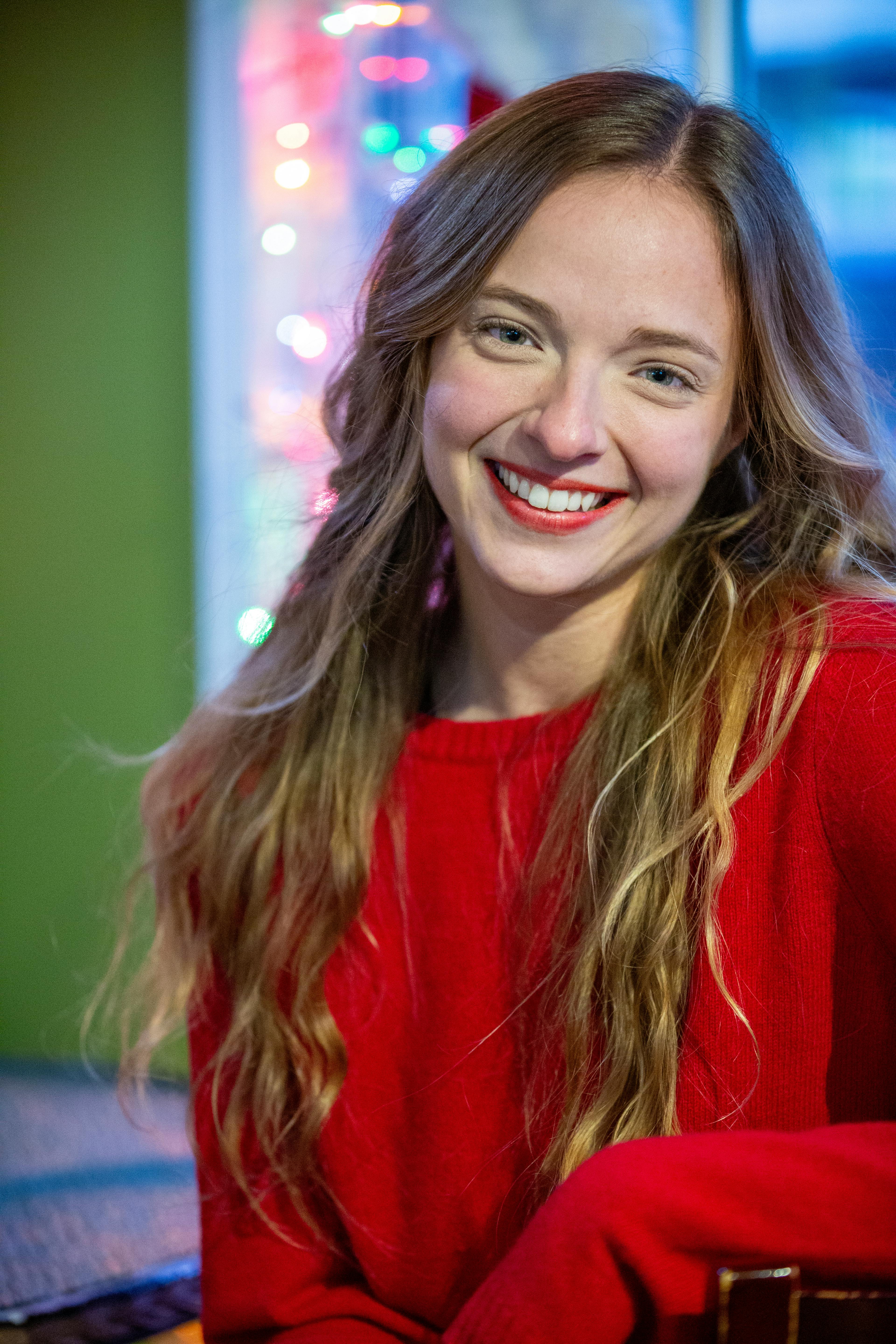 Smiling woman in red sweater with holiday lights background indoors.