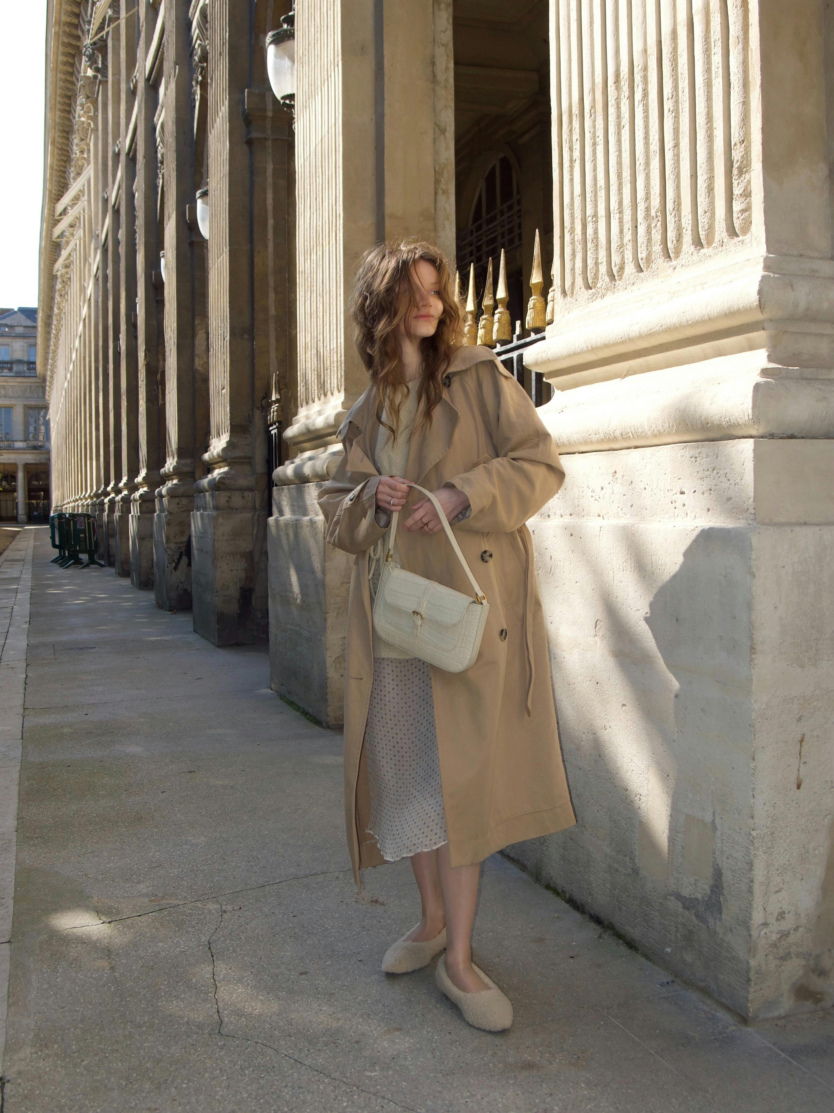 Stylish woman in a trench coat poses by historic architecture, embracing modern fashion.