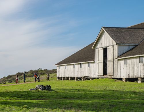 Free A rustic barn sits in a lush green landscape under a blue sky, with people enjoying the outdoors. Stock Photo