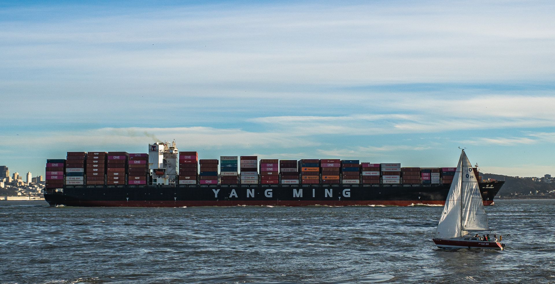 Container ship and sailboat navigating calm waters under a clear sky.