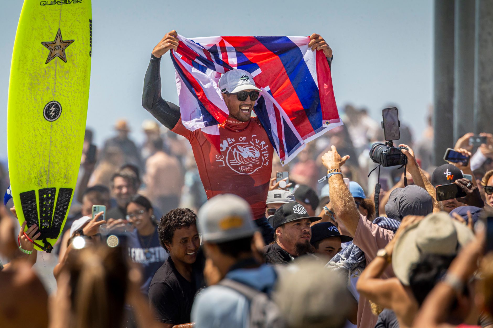 Surfer celebrates victory holding a vibrant flag, surrounded by a cheering crowd.