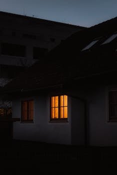 A cozy warm light shines through the window of a house at dusk, creating a peaceful urban scene.