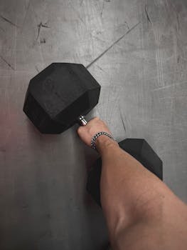 Close-up of arm lifting hexagonal dumbbell on gym floor, symbolizing strength and fitness.