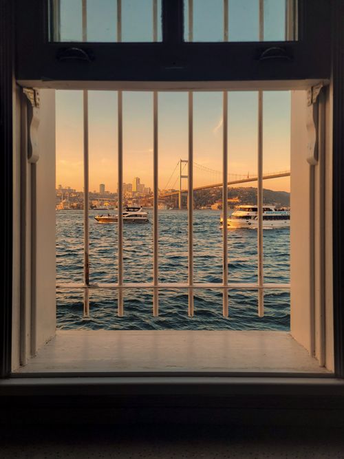 Free Boats pass under Istanbul's Bosphorus Bridge at sunset, seen through window bars. Stock Photo