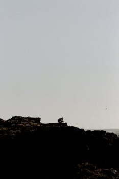 晴朗的天空下，一个人的剪影坐在崎岖的岩石上，这是白天拍摄的景象