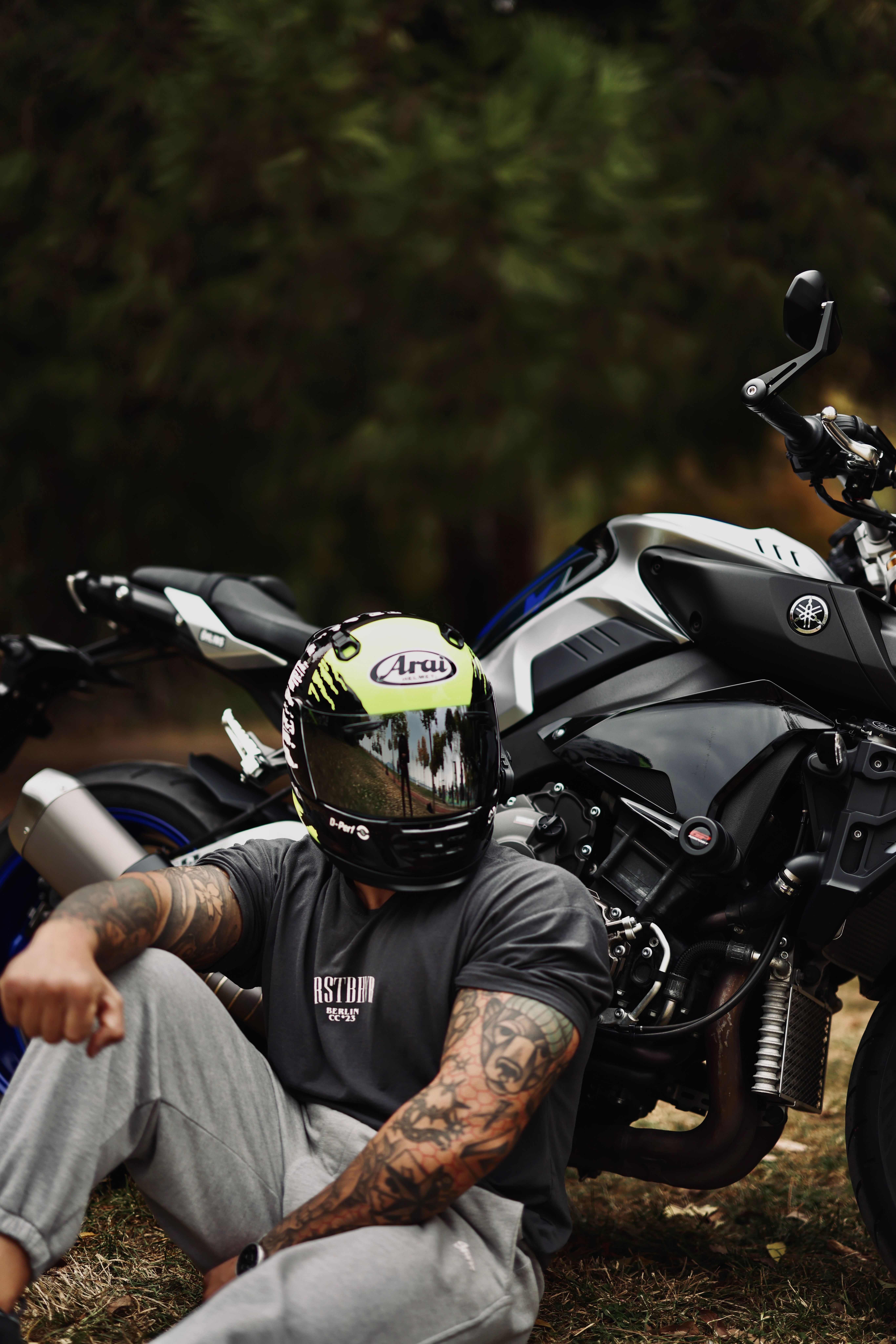Tattooed biker relaxing next to a motorcycle in a lush Istanbul park setting.