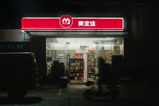 A dimly lit convenience store with a bright neon sign at night, featuring outdoor seating.