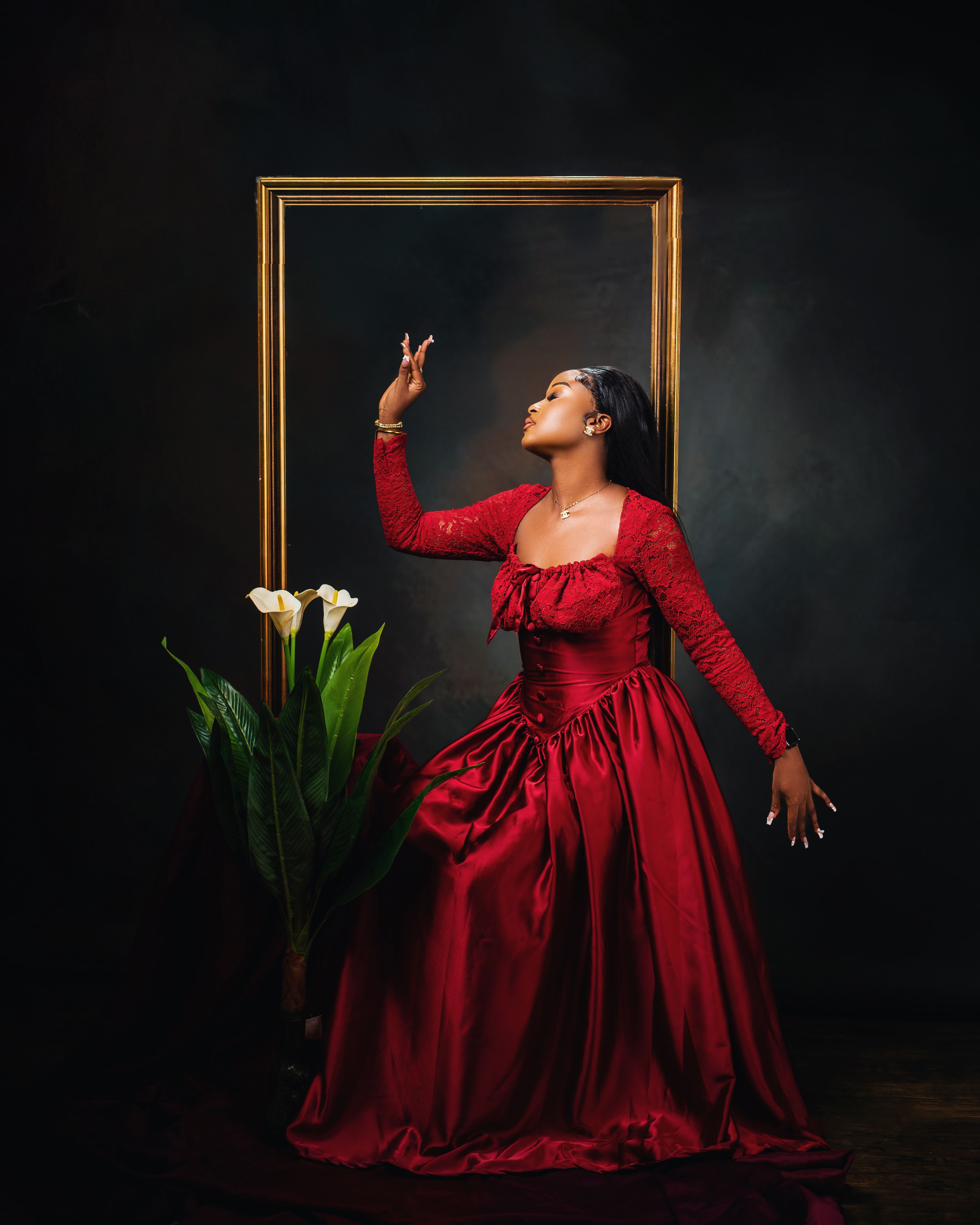 A graceful woman in a red gown poses beside a golden frame and white flowers.