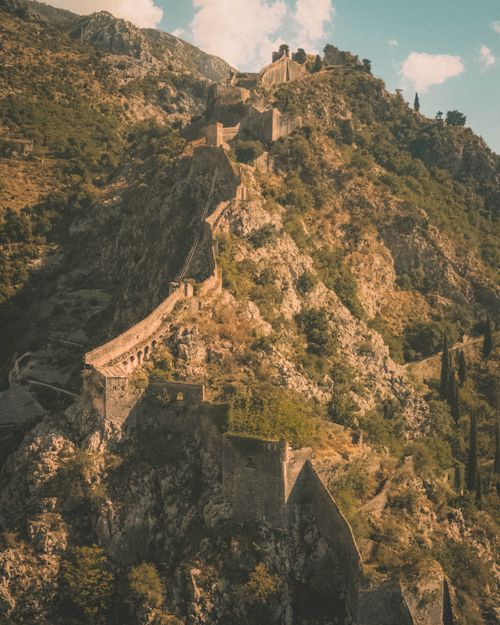 Free Aerial view of the dramatic Kotor Fort amidst Montenegro's rugged landscape under a warm sun. Stock Photo