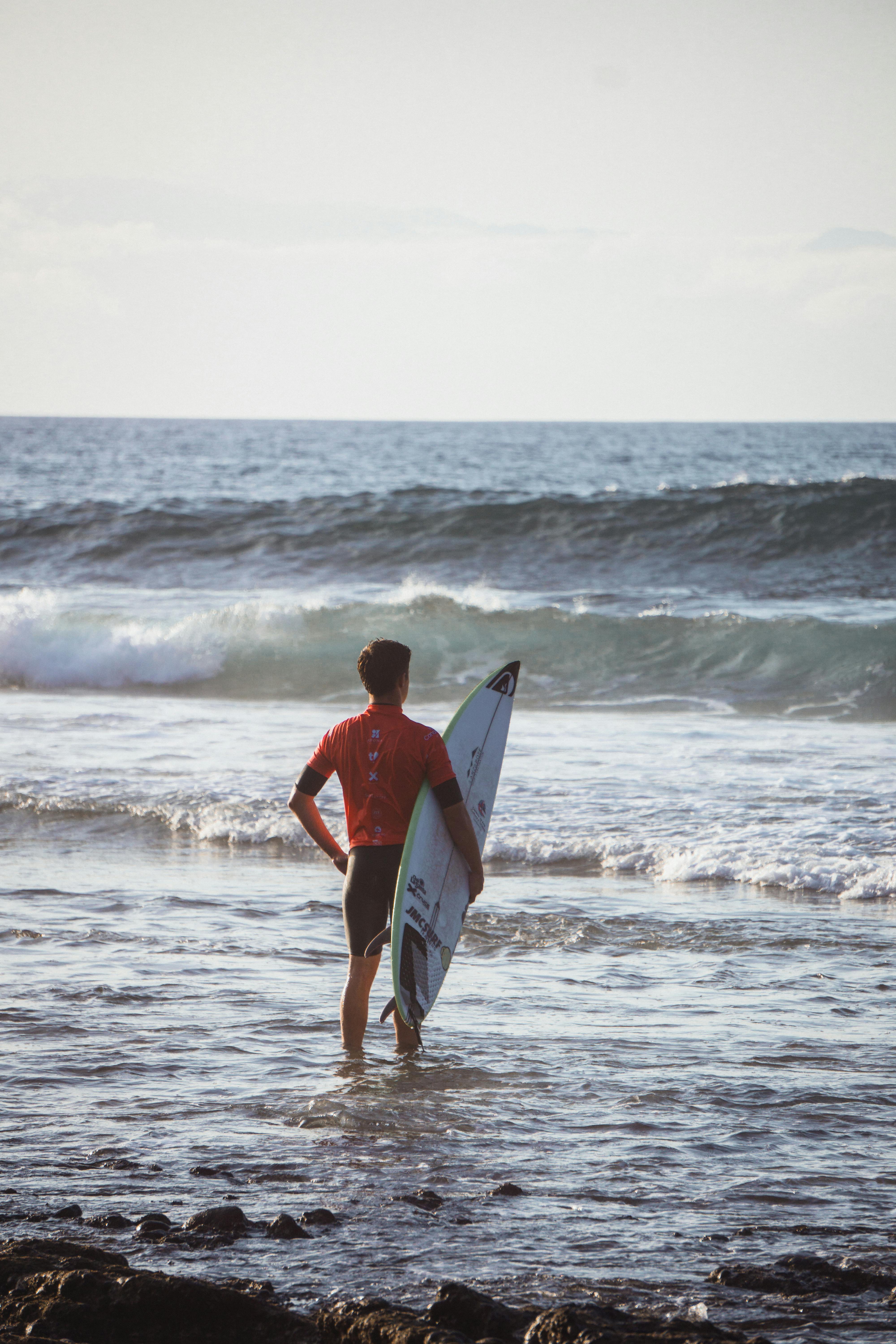 A young male surfer stands on the beach with his surfboard, gazing at the waves.