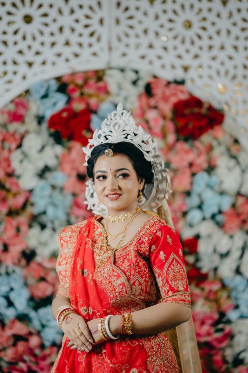 Free Charming bride adorned in vibrant red traditional dress against floral backdrop. Stock Photo