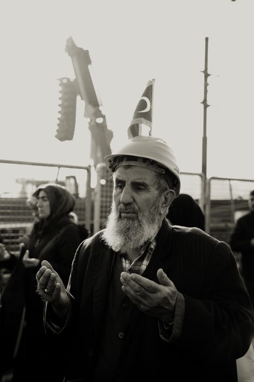 Free An elderly man prays at a construction site, wearing a hard hat with flag symbol, under a muted sky. Stock Photo