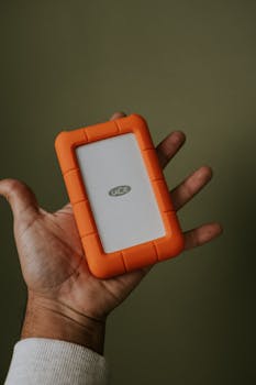 Close-up view of a hand holding an orange portable LaCie hard drive.