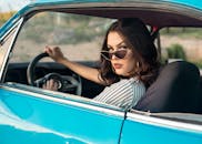 一位戴着太阳镜的妇女，在一个晴天驾驶着一辆老式蓝色汽车，散发着风格与自信