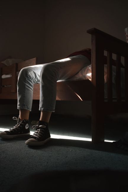 A person lies on a bed wearing jeans and sneakers, enveloped in shadowy light.