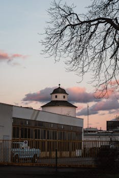 An industrial building framed by leafless trees at sunset, with a peaceful pink sky.