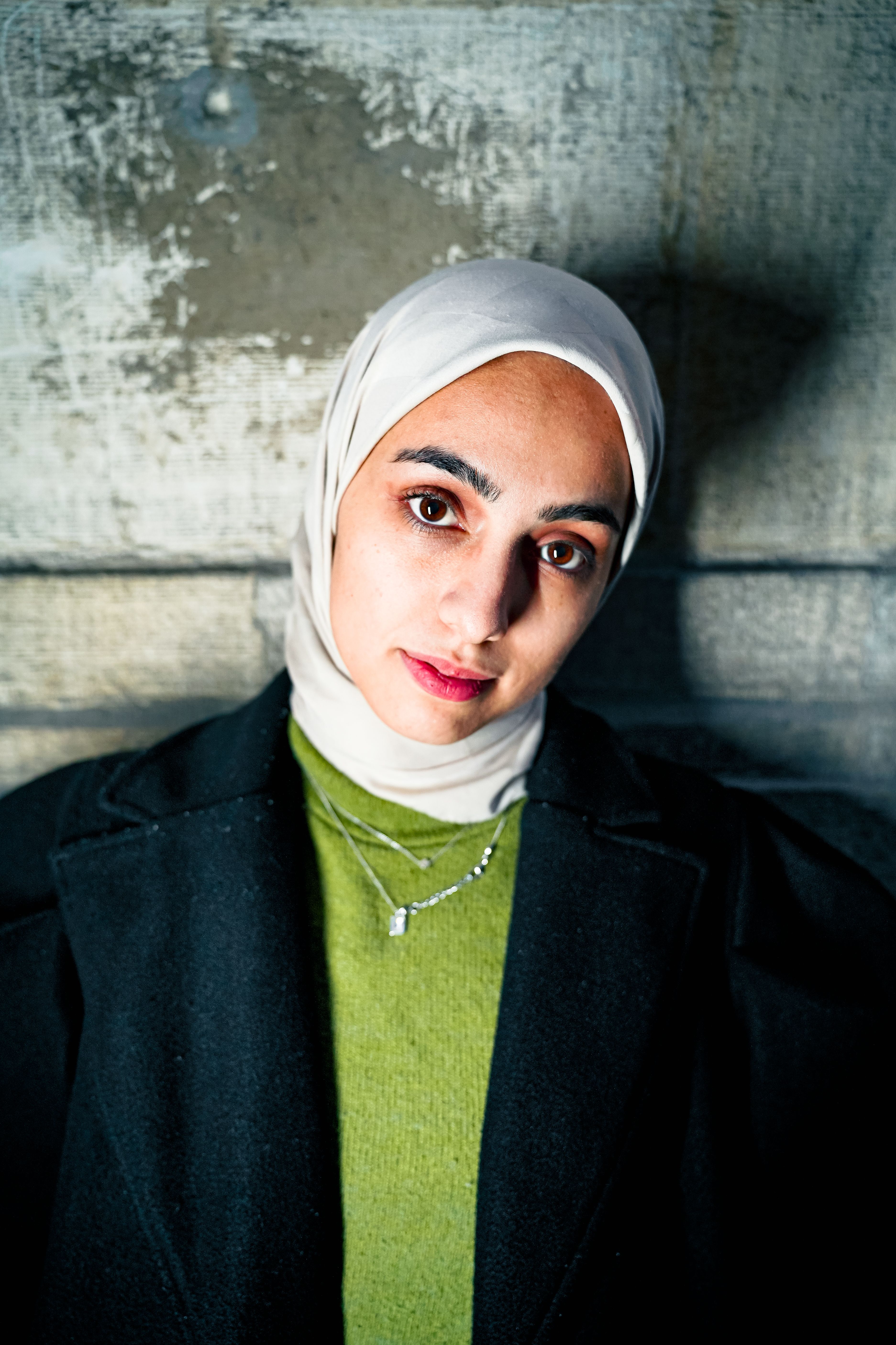 A woman wearing a hijab and coat stands against a textured wall, looking intently at the camera.