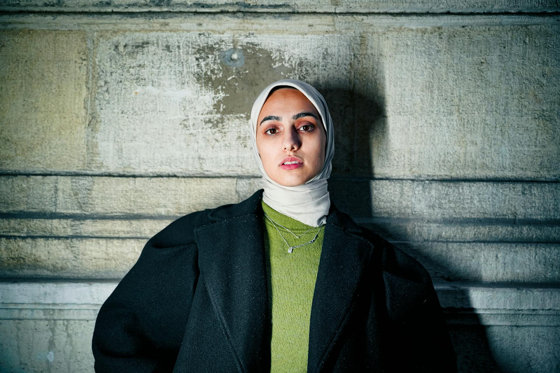 Portrait of a woman in hijab standing against a textured stone wall, exuding confidence and poise.