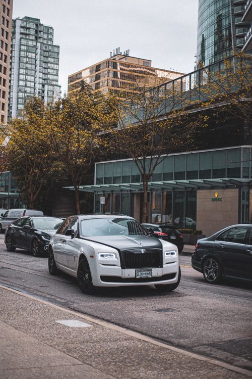 Free White Rolls Royce cruising through a city street with modern architecture and autumn foliage. Stock Photo
