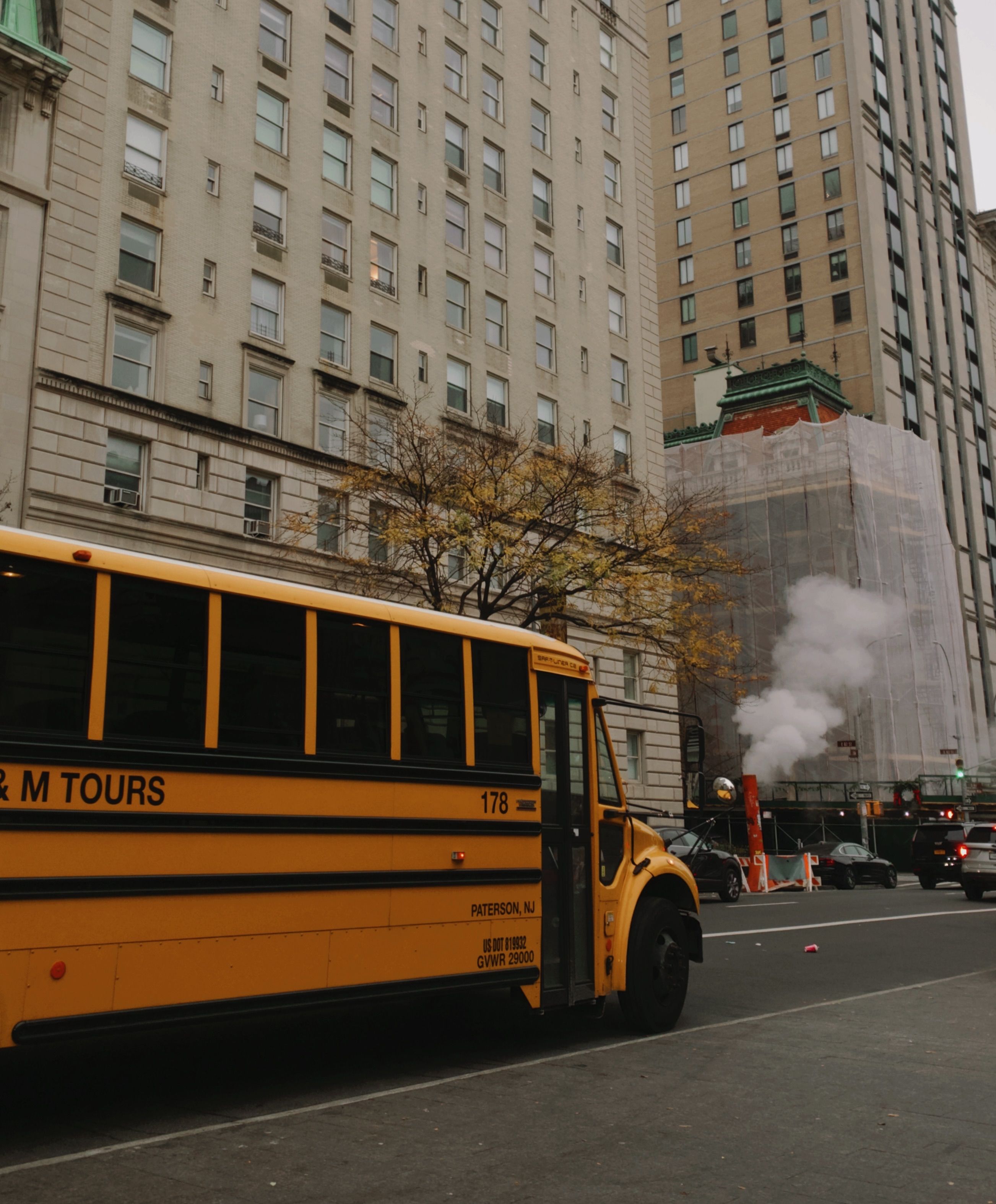 A yellow school bus parked in front of city buildings during autumn, with steam rising nearby.