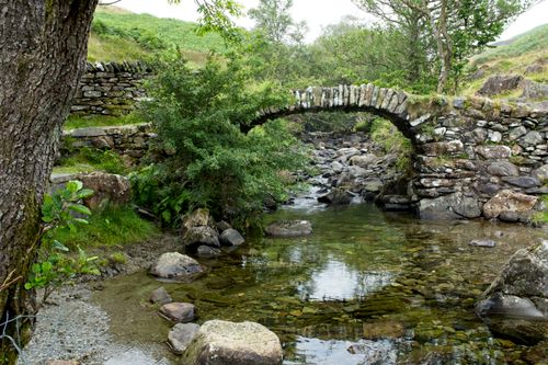 Free A serene stone arched bridge over a reflective stream in a lush countryside setting. Stock Photo