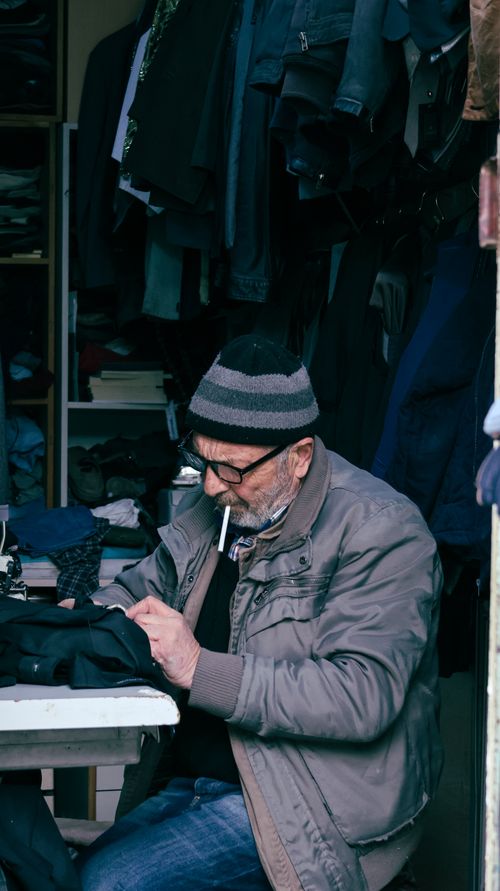Free An elderly man tailoring clothes in a small, dimly lit workshop, wearing glasses and a hat. Stock Photo
