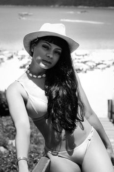 A woman in a bikini and sun hat on a bustling beach in black and white.