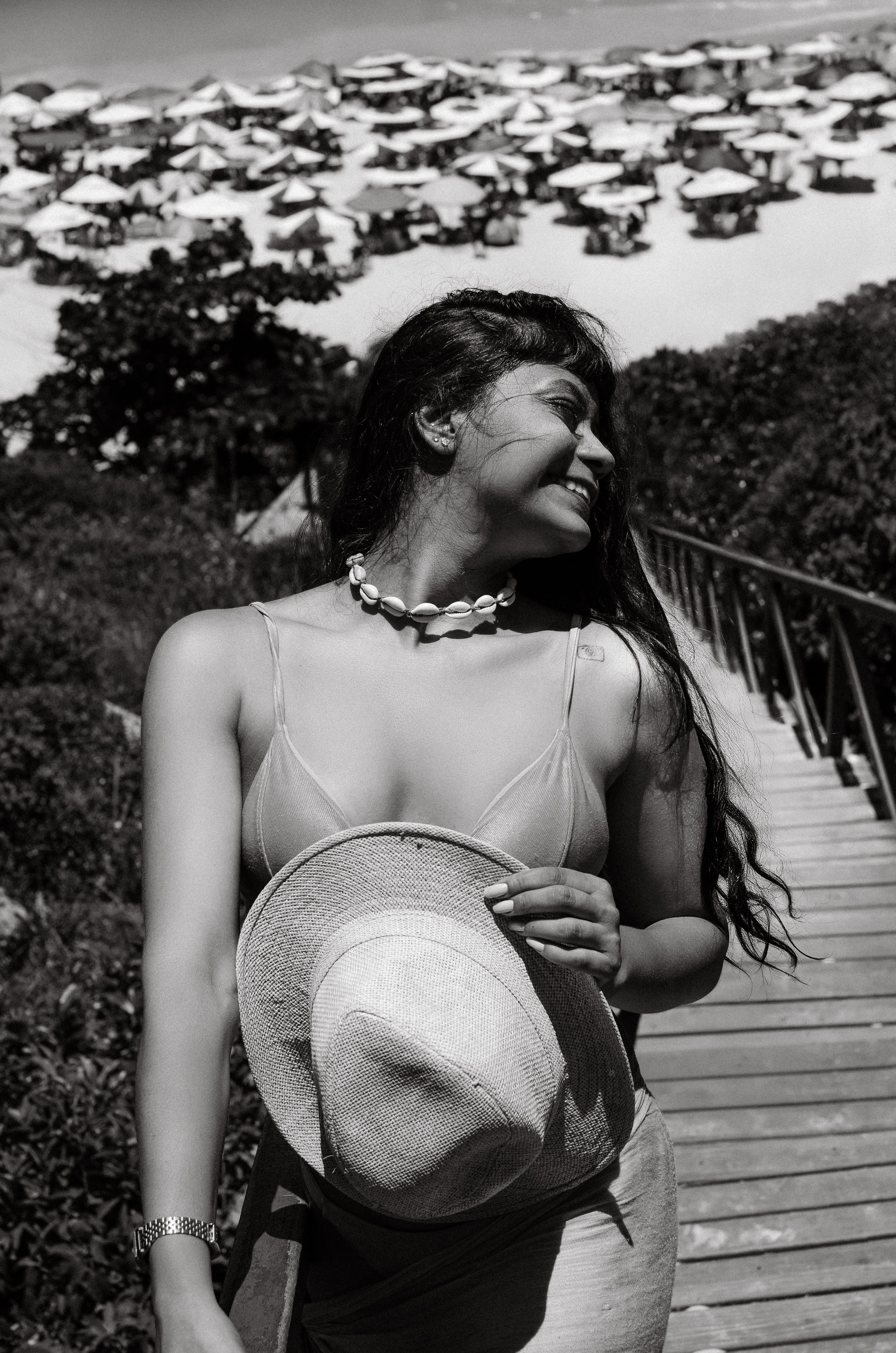 Black and white photo of a smiling woman on a beach boardwalk, holding a hat.