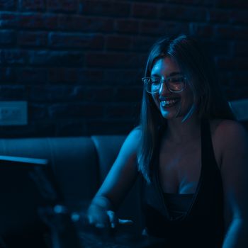 A young woman wearing glasses smiles while working on her laptop in a dimly lit room with a brick wall background.