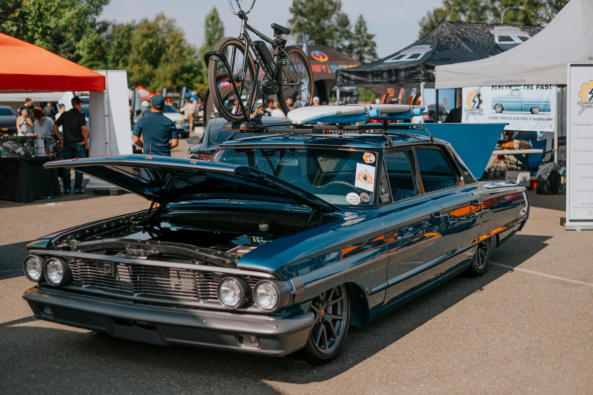 Vintage car with an open hood and bicycle on top at an outdoor auto event.