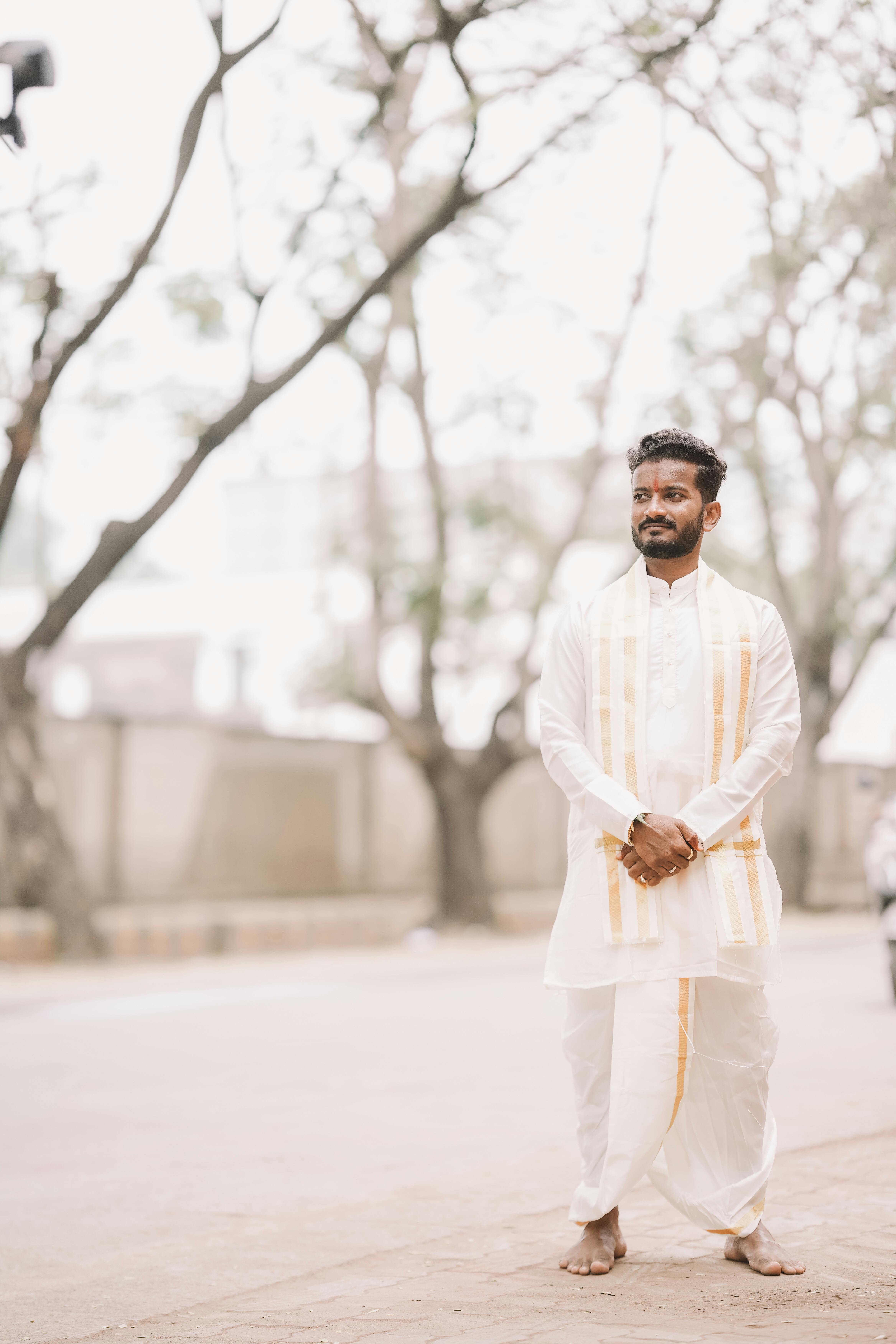 A man in traditional white and gold attire standing outdoors on a bright day.