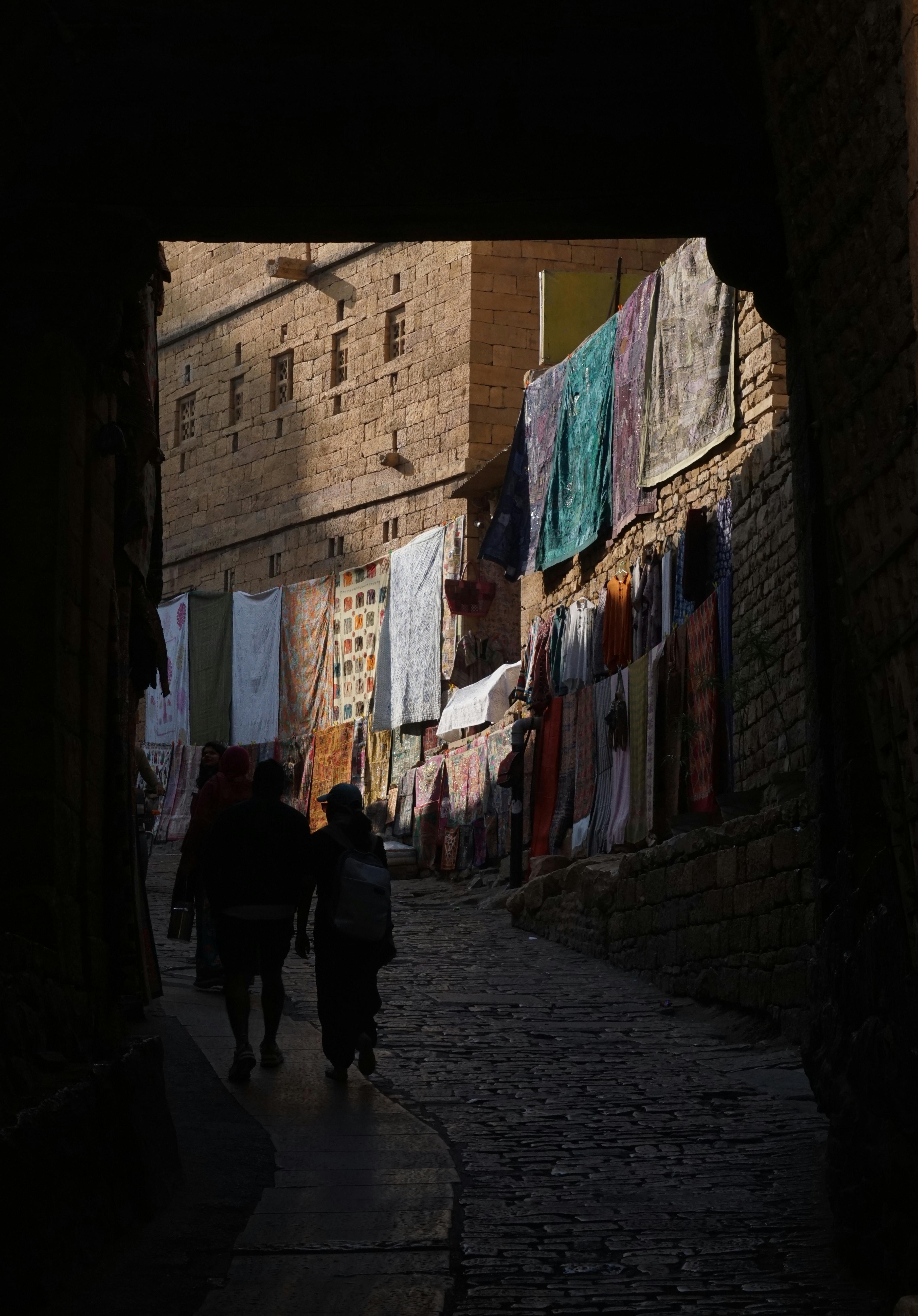 Silhouetted figures walking in a narrow alley with hanging textiles in Jaisalmer, Rajasthan, India.
