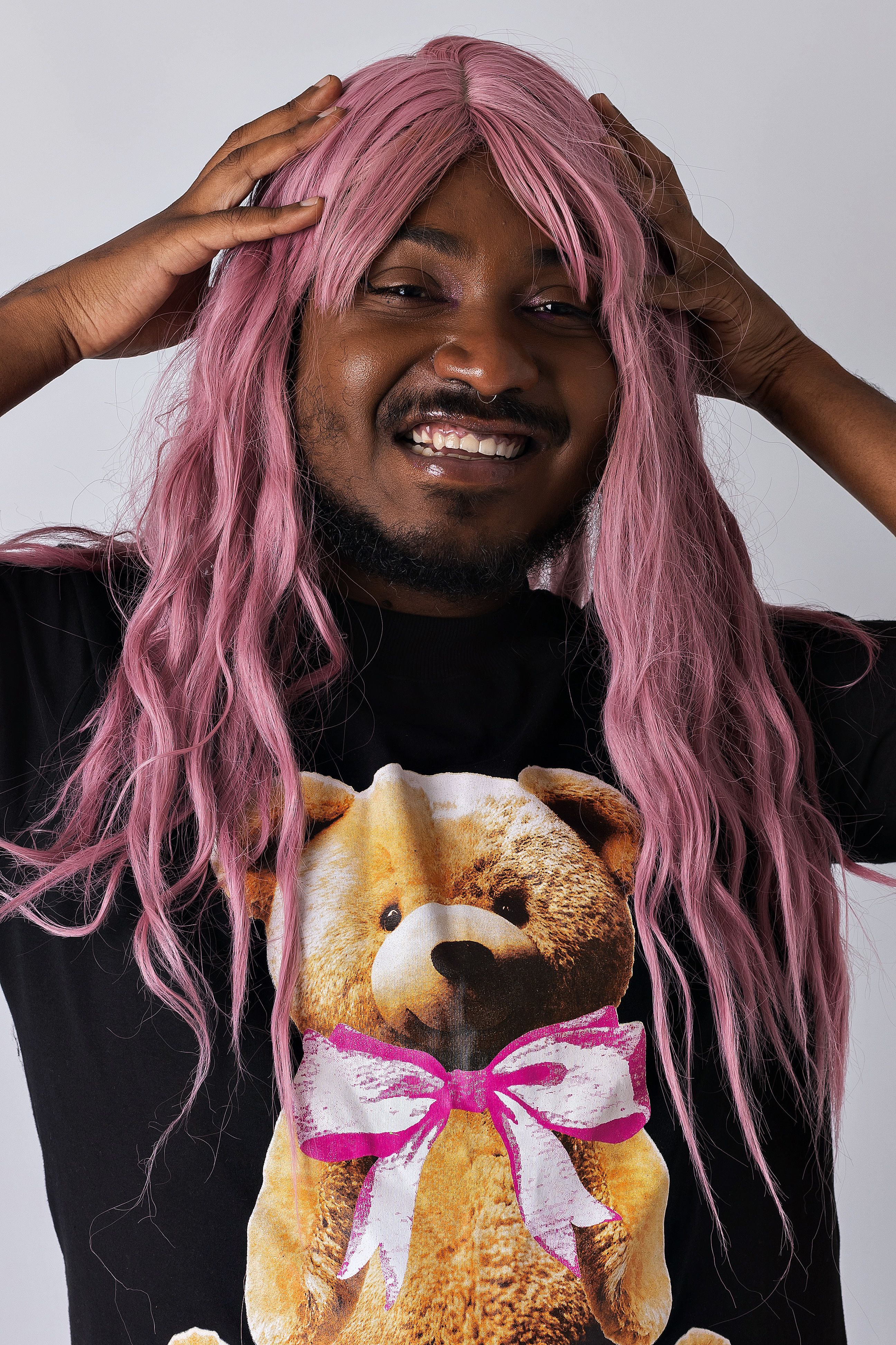 A joyful portrait of a person wearing a pink wig and a teddy bear shirt, smiling broadly.