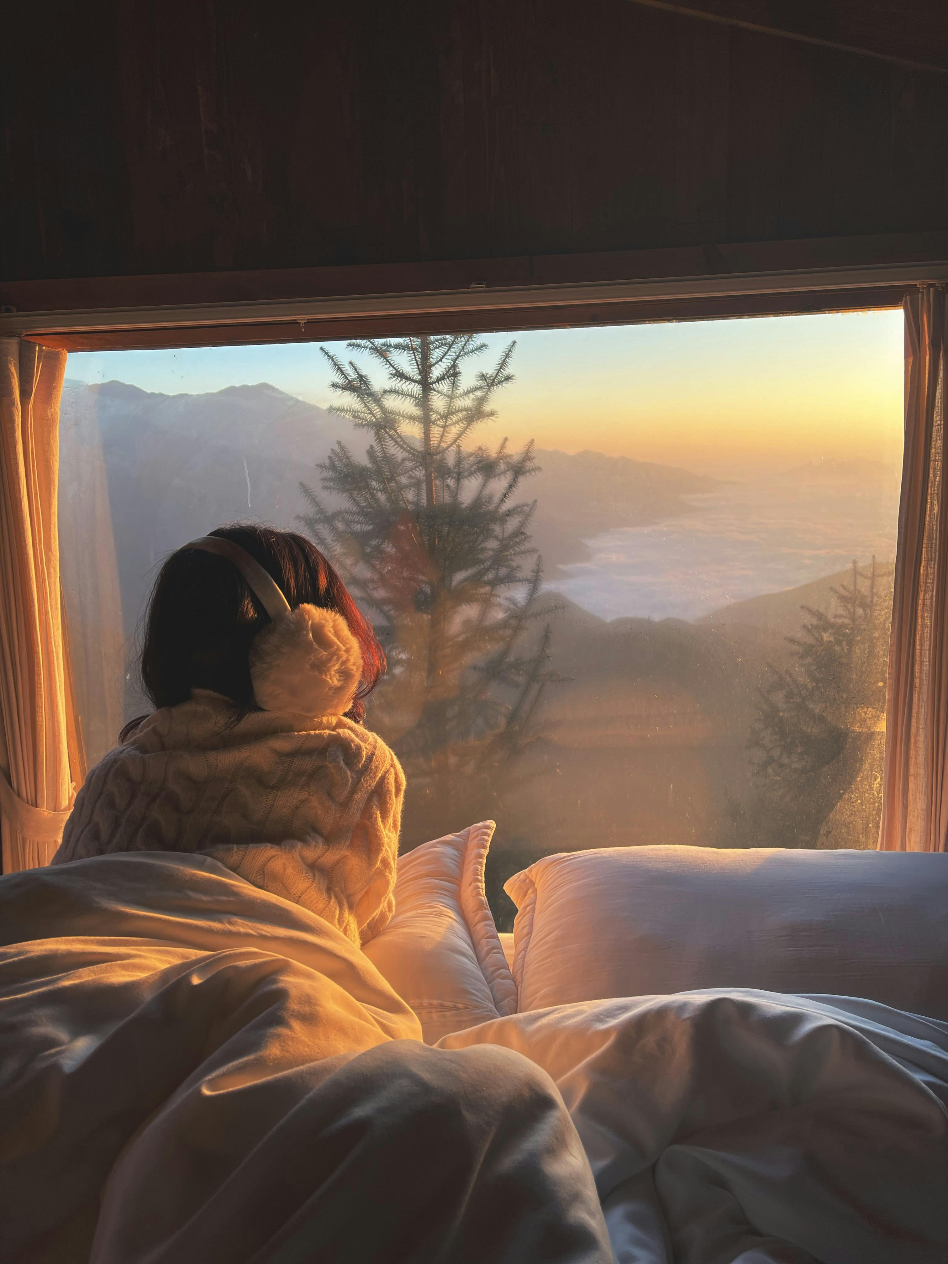 A person enjoys a cozy morning view of mountains and sunrise from bed.