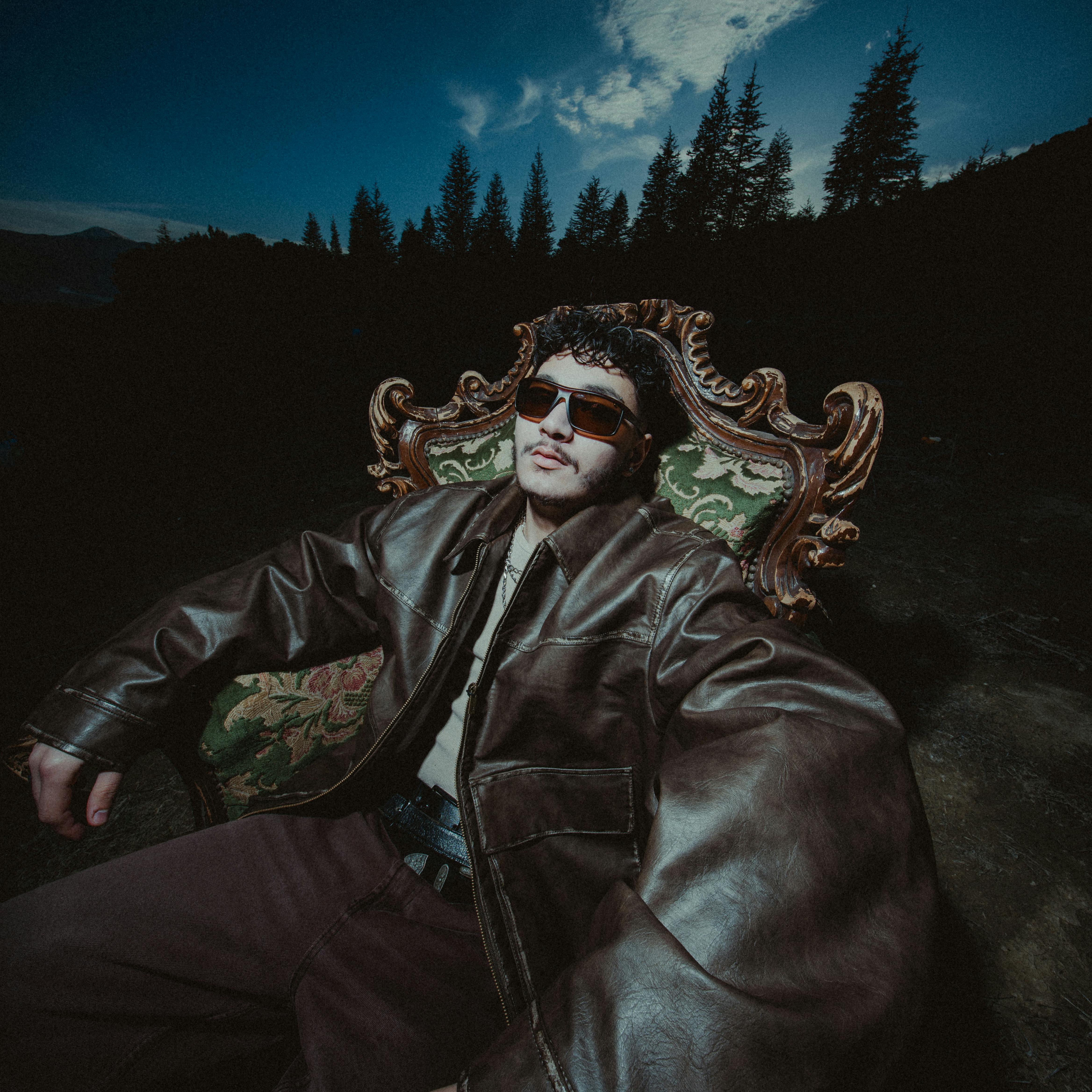 Young man in sunglasses and leather jacket seated outdoors at dusk with trees in the background.
