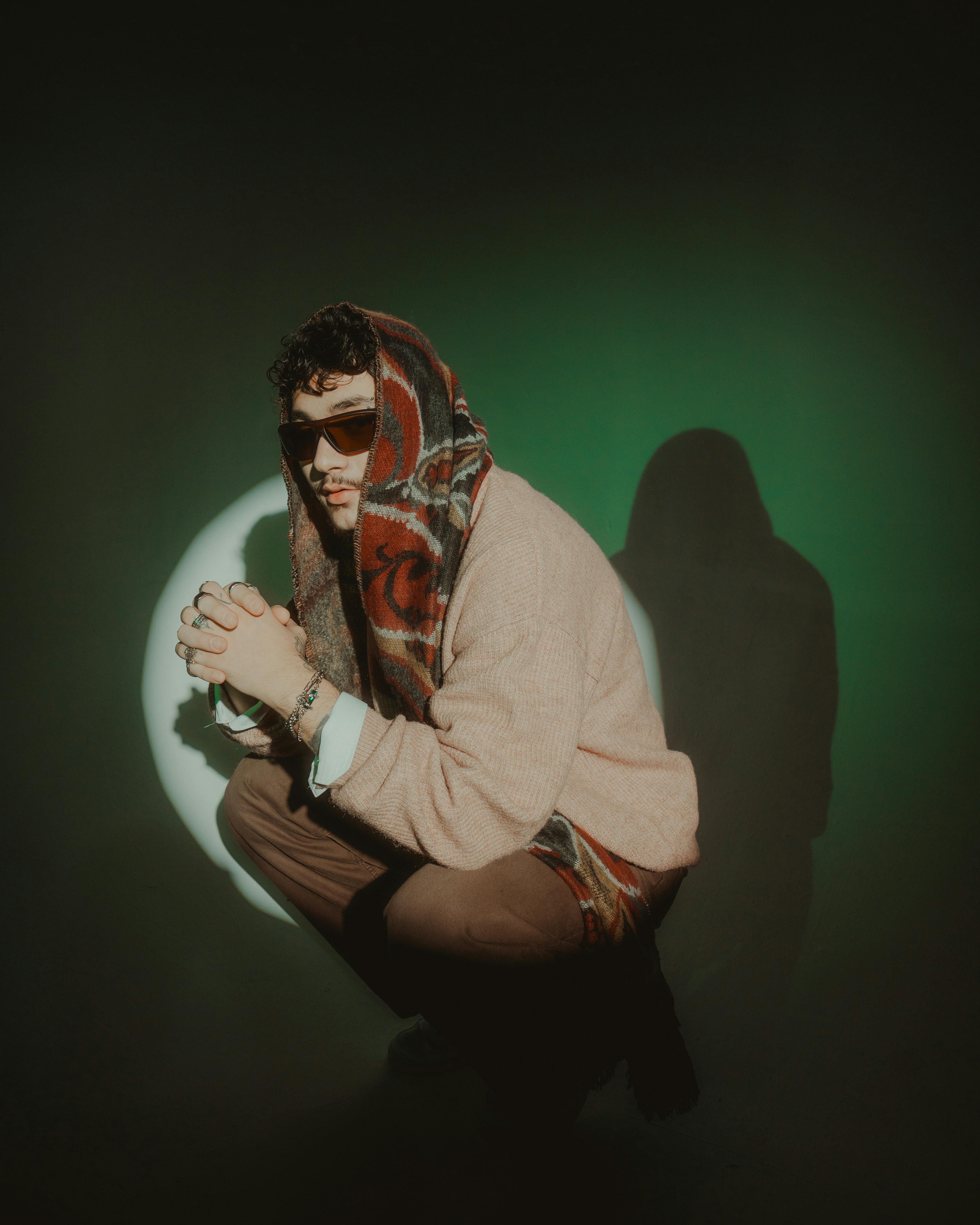 A fashionable man in sunglasses and a colorful scarf seated thoughtfully against a moody backdrop.
