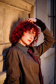 Fashionable woman with red curly hair poses against city wall, exuding confidence and style.