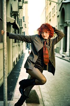A woman with curly red hair in a suit striking a confident pose in a narrow street.