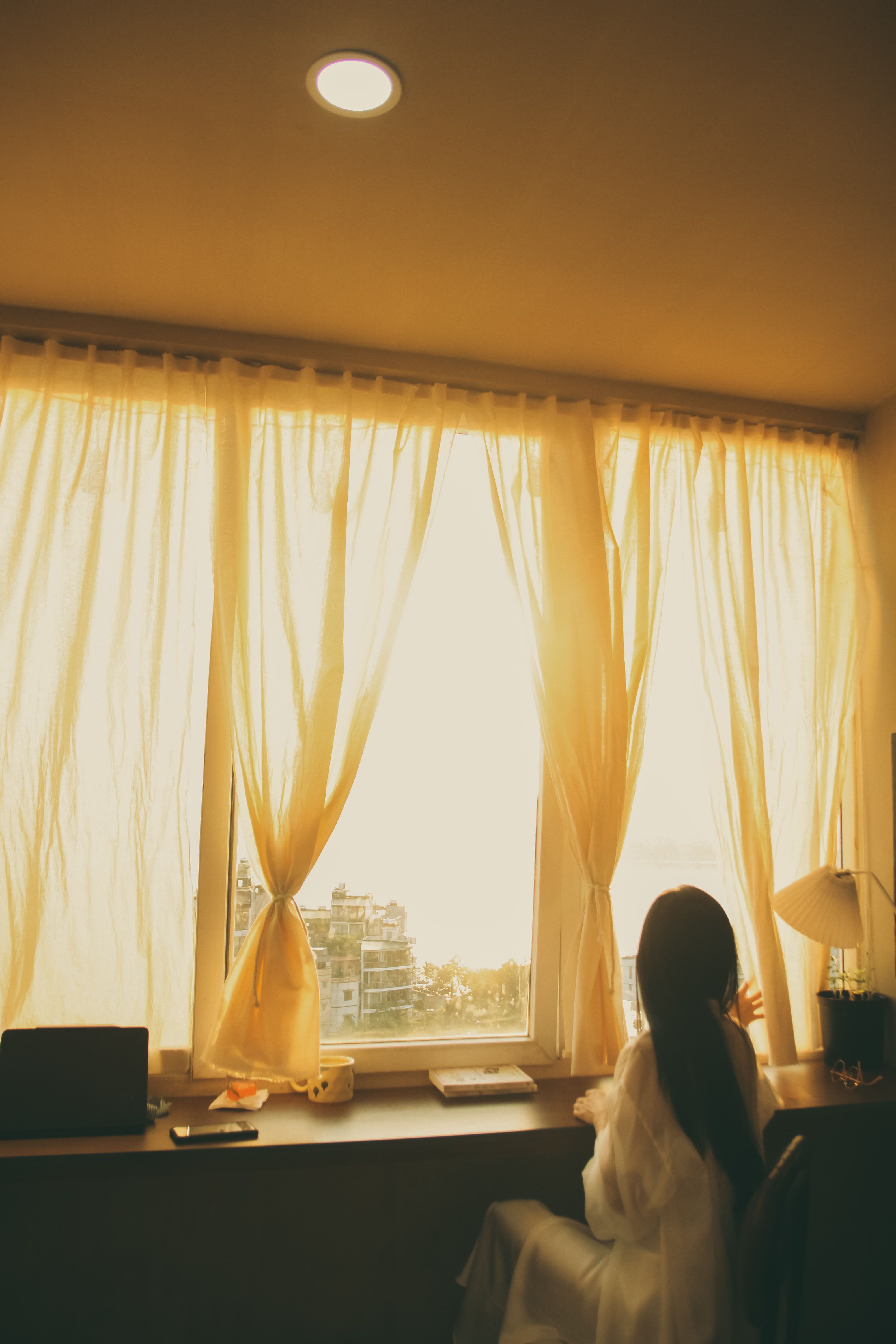 Silhouette of a woman looking out a sunlit window with soft curtains on a calm day inside.