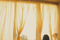 Silhouette of a woman looking out a sunlit window with soft curtains on a calm day inside.