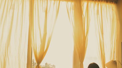 Silhouette of a woman looking out a sunlit window with soft curtains on a calm day inside.