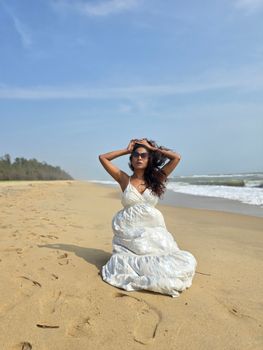 A woman in a white dress poses on the sandy shores of Puducherry's beach under a clear blue sky.