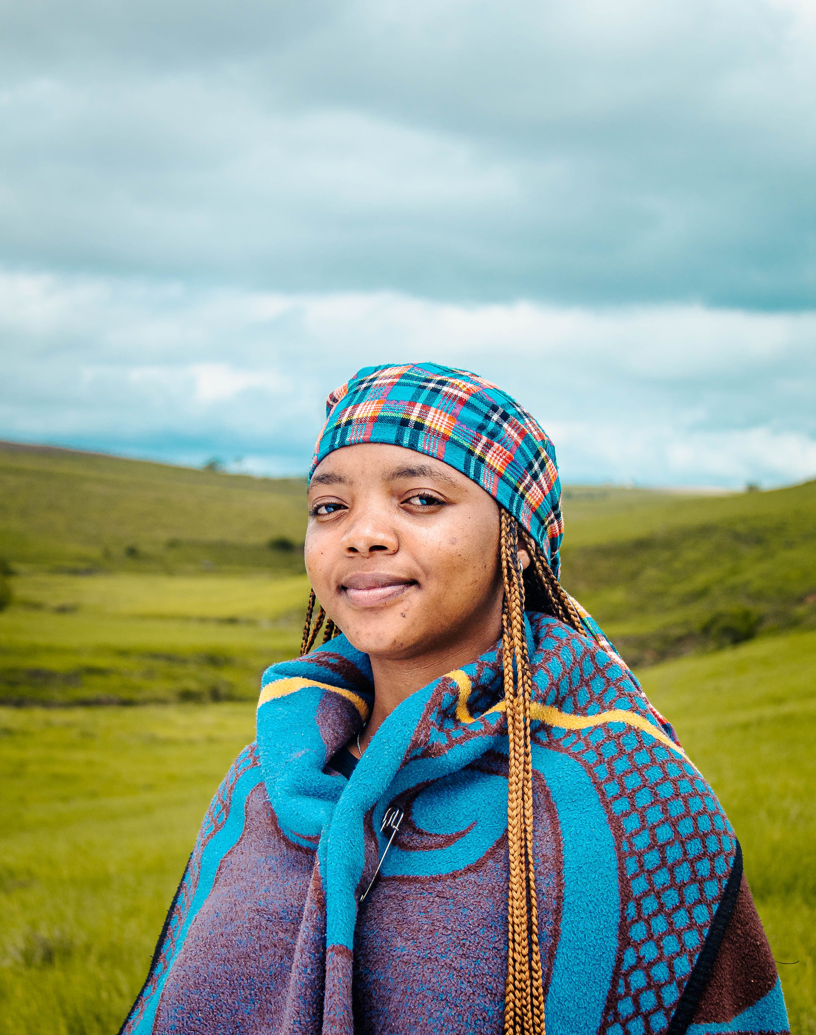 A woman wearing colorful traditional attire, standing in a lush green field in South Africa.