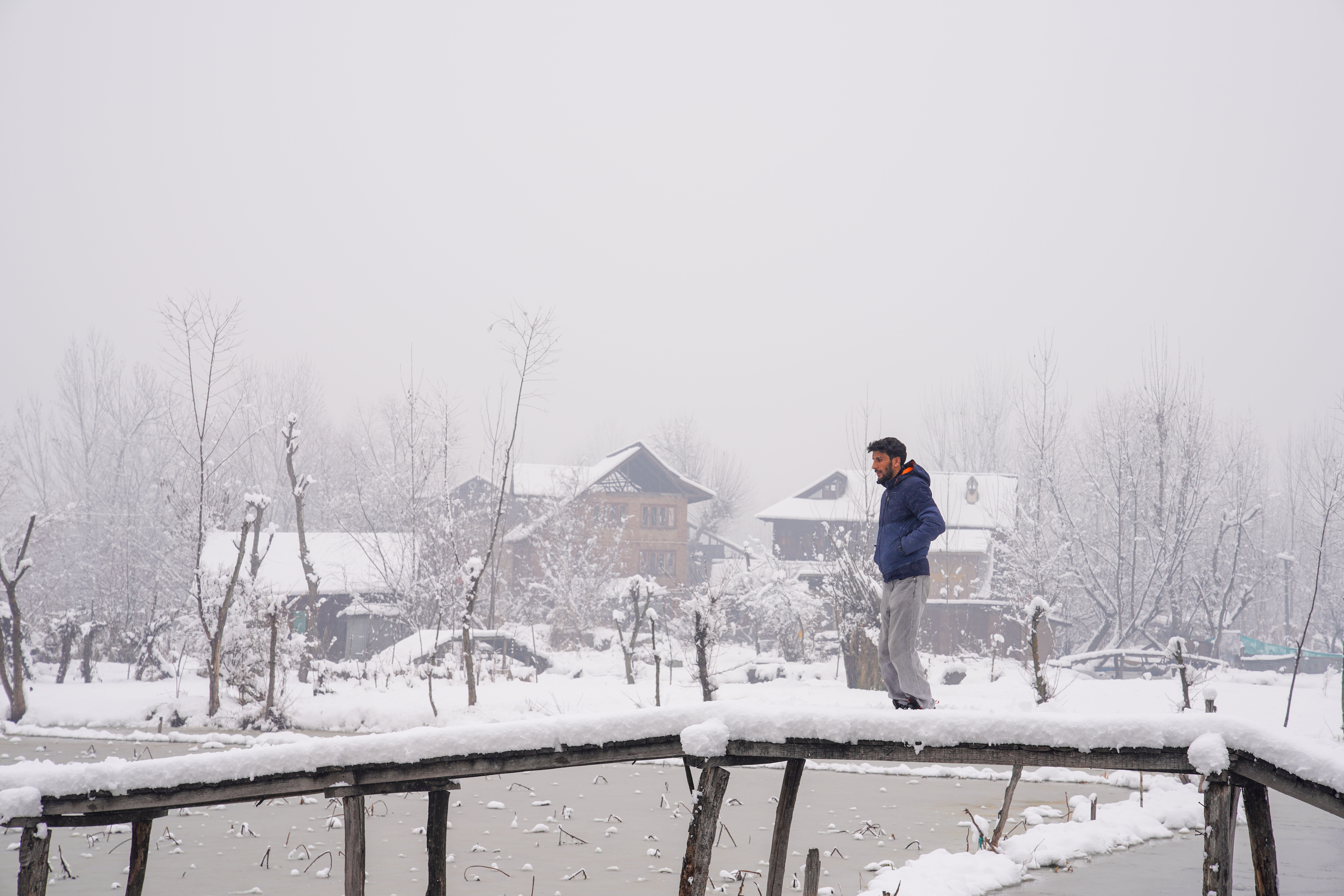 A serene winter scene featuring a man walking on a snowy bridge in a Kashmir village.