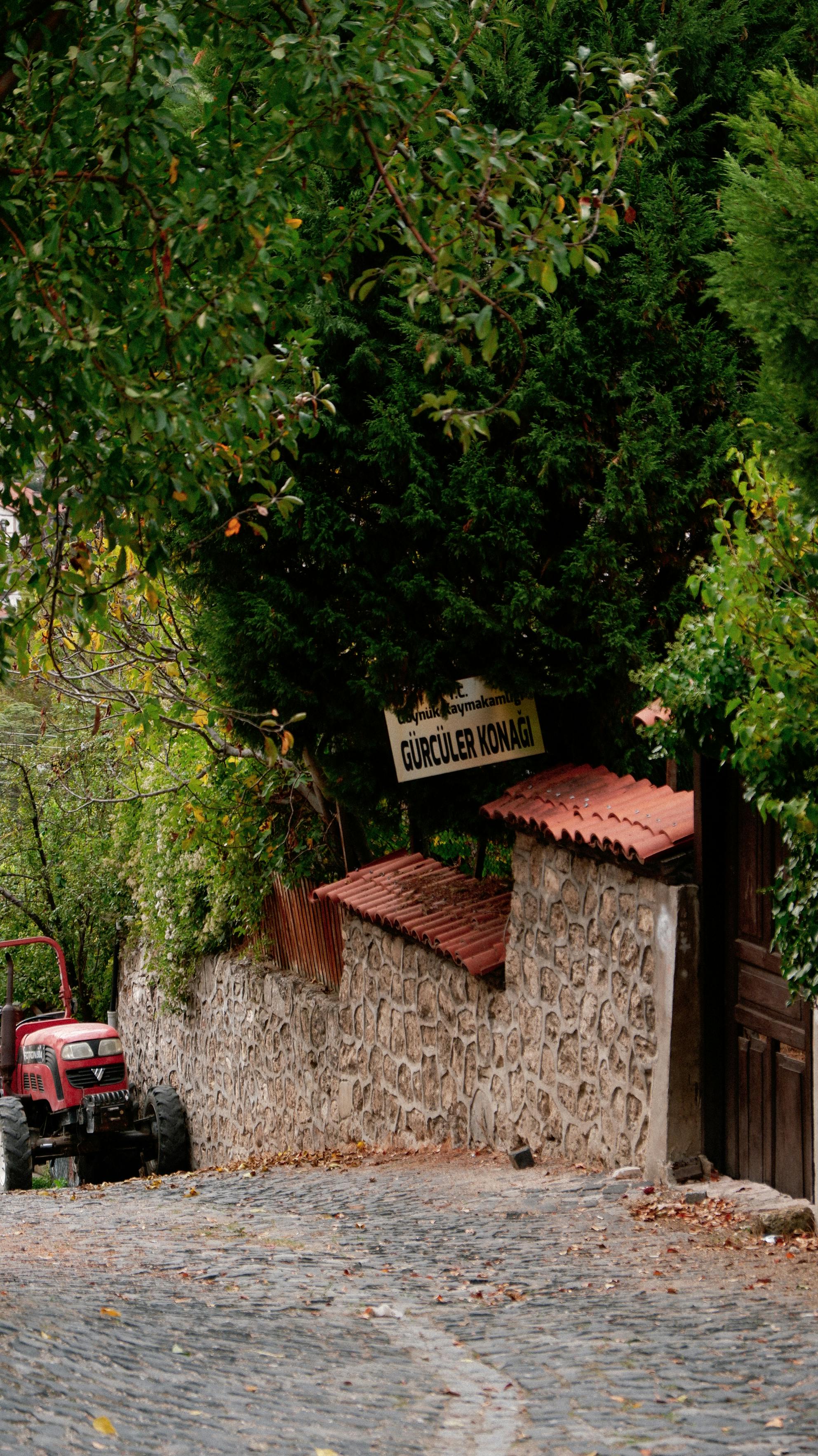 A quaint cobblestone street lined with trees and a red tractor parked along a stone wall in a charming village setting.