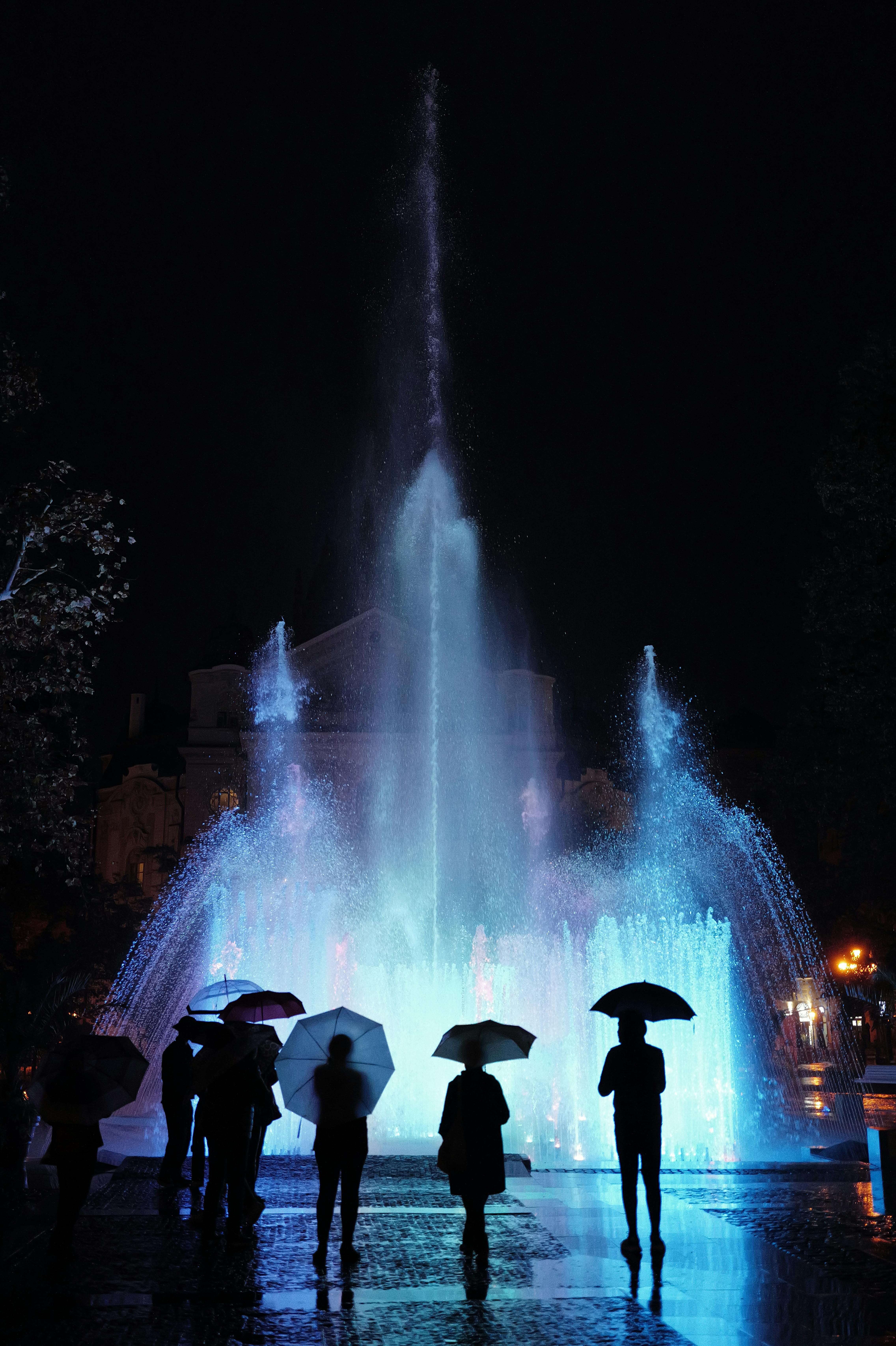 Night scene of people holding umbrellas by a glowing blue fountain.