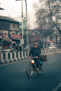 A man bikes along a road in Srinagar, surrounded by winter scenery and urban architecture.