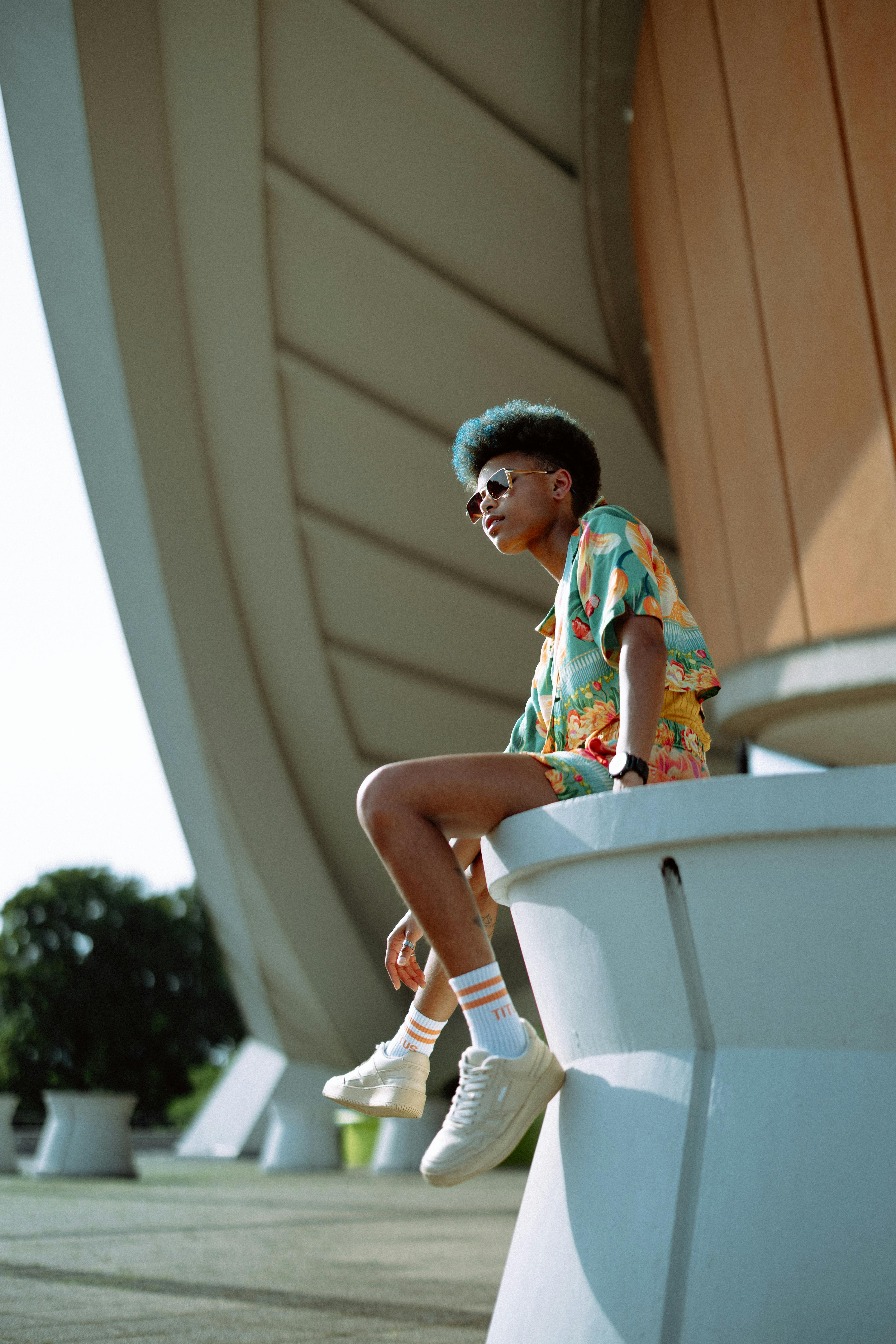 Gratis Un individuo elegante, vestito con vivaci abiti estivi, si gode una giornata di sole all'esterno di una struttura moderna. Foto a disposizione